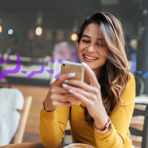RCS Messaging image Photoillustration of a woman holding an iPhone in both of her hands and smiling at the screen; she has long, wavy brown hair and light coffee-colored skin and she's wearing coral lipstick; on top of the photo, there's an overlay graphic of message bubbles