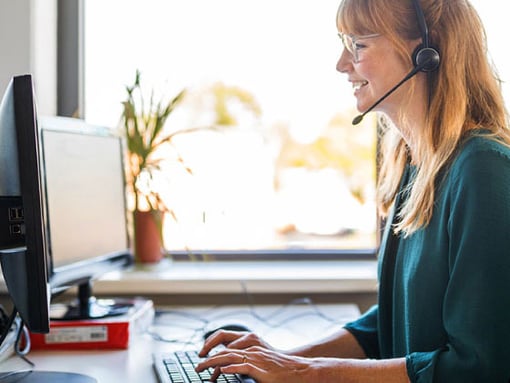 UC Cloud Call Center Article Header Photo of a smiling female call center agent talking to a customer on her headset as she types notes on her computer.